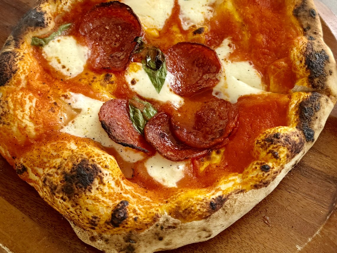 A home-baked Neapolitan pizza with salami, fior di latte and basil, its puffed rim charred in spots, on a wooden board